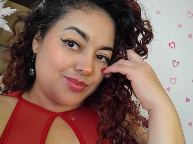 Lalitzfet a les cheveux roux-brunâtres bouclés, elle porte un haut rouge transparent, avec du rouge à lèvres et de l'eye-liner, et elle touche sa joue avec une ongle rouge, tout en souriant légèrement..