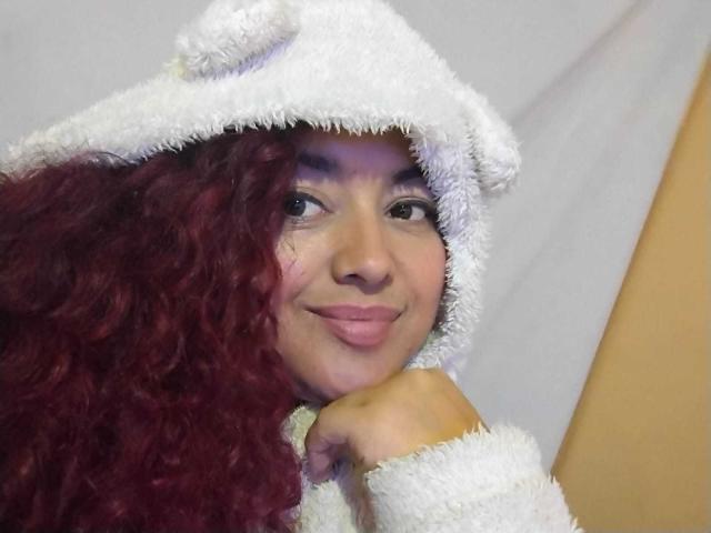 Lalitzfet with curly red hair, wearing a fluffy white bear hat, smiling with puffy pink lips, resting chin on hand, soft facial expression, light brown skin.