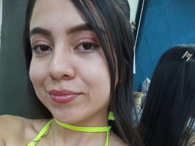 JessyLenon has shiny brown eyes, pink eyeshadow, glossy pink lips, and a yellow choker. She's smiling with black hair, a hairpin, and a mirror reflection.