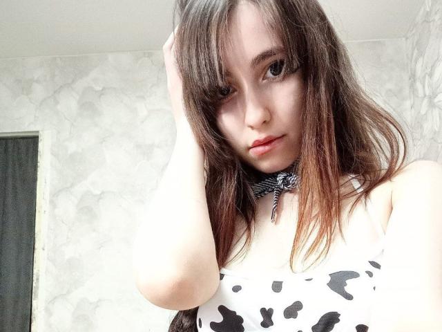 SavyLoly with long brown hair, wearing a cow print top, and a black-and-white choker, touching her hair, showing a cute, slightly sultry face, and small tits.