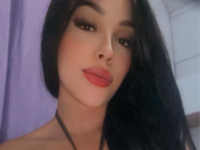 Camilla Carioca hat eine glatte, gebräunte Haut, volle, rote Lippen, langes, schwarzes Haar und dunkle Augenbrauen. Sie trägt ein schwarzes Top mit Halstiefe, hat einen leicht verführerischen Blick und trägt dezentes Make-up..