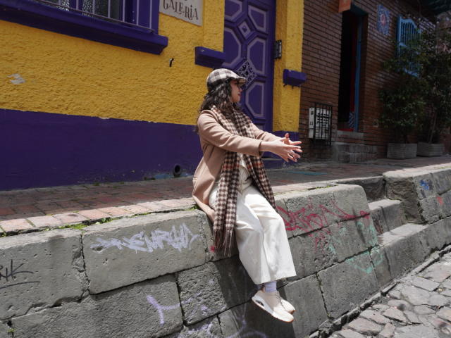 ElizaCanterville, sitting on a gray stone wall, wears a beige coat, white pants, brown checkered scarf, and white shoes, with a purple and yellow building behind her.