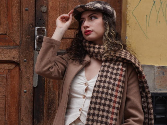 ElizaCanterville with curly brown hair, brown coat, white vest, houndstooth scarf, and flat cap, touching her hat, showing red lipstick and a slight smile.