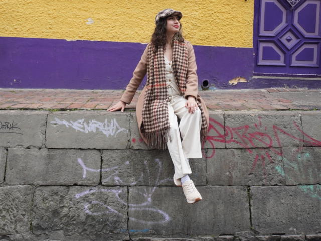ElizaCanterville sitting on a brick wall, wearing a tan coat, checkered scarf, white pants, white sneakers, and a flat cap, with a yellow and purple wall behind.
