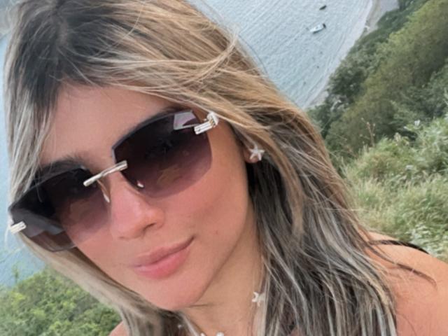 SashaPolet with blonde and brown hair, wearing dark sunglasses and star earrings, has a pouty face and is looking at the camera. Her tits are partially visible.