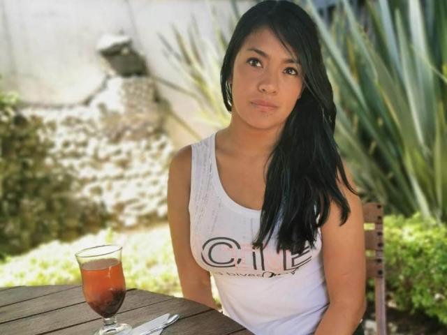 ArianithaXox with long black hair, medium brown skin, and cute face, wearing a white tank top, sitting at a wooden table with a glass of iced tea.