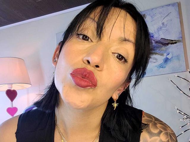 MadissonMayer with black hair, puffy red lips, small nose, tattoos on shoulder, black sleeve, gold earrings, and necklace, making a puckering face in a photo.