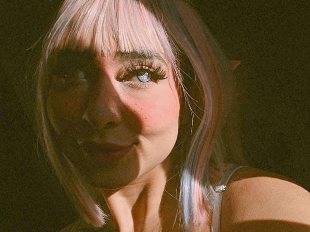 BlueSkarlet has pinkish-white hair, elf-like ears, and long, thick eyelashes. Her light skin and blue eyes are highlighted by sunlit shadows on her face.
