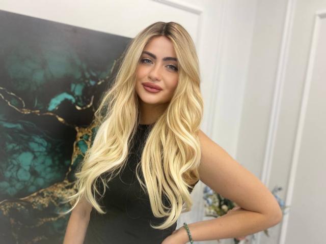 LeslieMineg has long blonde wavy hair, blue eyes, and is wearing a sleeveless black top, showing her tits slightly, with a green bracelet on her right wrist.