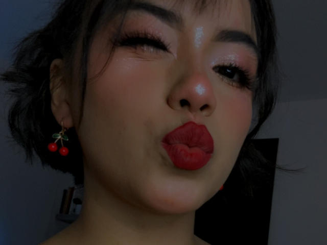 Kathie, con la piel brillante, lápiz labial rojo y aretes de cereza, frunciendo los labios y guiñando un ojo. Tiene el cabello oscuro, cejas pobladas y pestañas largas. Su rostro está centrado..
