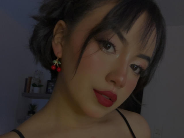 Kathie tiene el cabello oscuro recogido de forma descuidada, lápiz labial rojo y aretes rojos en forma de cereza, con un maquillaje sutil y delineador de ojos negro, mirando ligeramente hacia un lado en una habitación con poca luz..