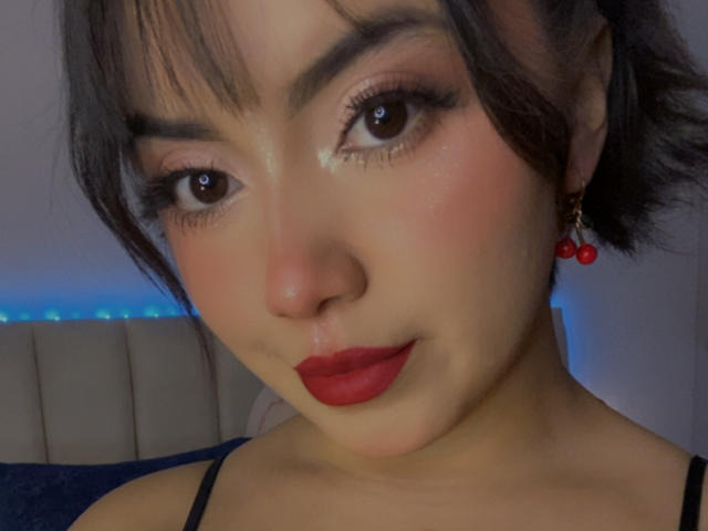Kathies has shiny brown eyes, red lipstick, and short black hair with bangs. She's wearing red cherry earrings and a black strap top, showing a cute, sexy face.