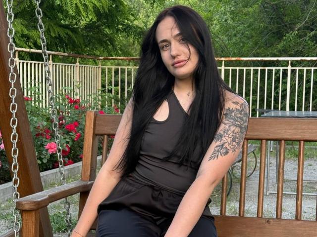 SaraFlame met lang zwart haar, een zwarte tanktop en zwarte broek, zittend op een houten schommel, met een tatoeage op haar rechterarm, die lichtelijk opzij kijkt en glimlacht..
