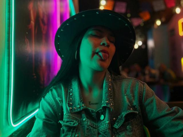 EmiliaCartersh met groen neonlicht op haar gezicht, tong uitgestoken, gekleed in een spijkerjasje met studs en een zwarte cowboyhoed, lang zwart haar, tatoeage onder haar kin, in een bar met weinig licht..