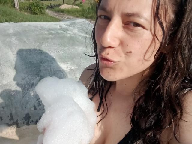 MilaWilde with wet, dark hair, pouty lips, and small moles on her face, holding a foamy white substance against her tits in a sunlit, outdoor setting.