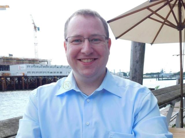Nilswinger24-hot, bald with glasses, wearing a light blue shirt, smiling at the camera, has a chubby face and is sitting outside under a big umbrella.