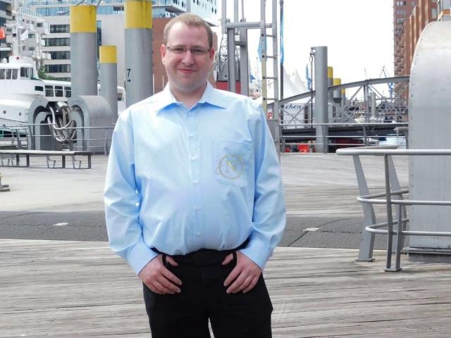 Nilswinger24-hot, a chubby dude with glasses, wearing a light blue shirt and black pants, standing on a wooden dock, hands on hips, smiling at the camera.