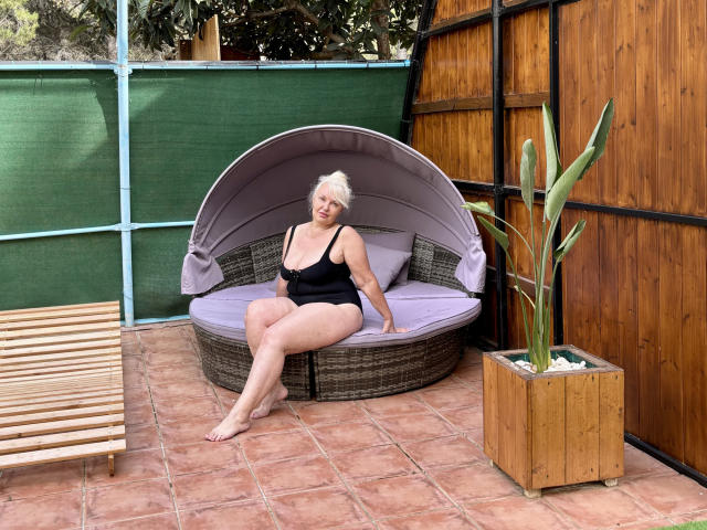 KamaliaSim with white hair, black one-piece, sitting on a wicker chair with purple cushions, showing her ass, in a patio with wooden walls and a potted cactus.
