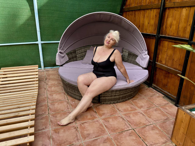 KamaliaSim, a chubby woman with short blonde hair, sits on a round wicker chair with a purple cushion, wearing a black one-piece, showing her big ass and small tits.