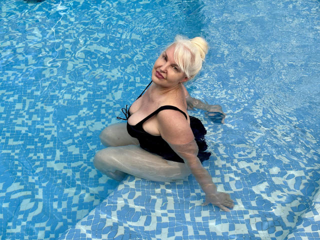 KamaliaSim, blonde, in blue-tiled pool, wearing black one-piece, sitting with ass in water, looking up, waterlines on tits and ass, arms resting on pool steps.