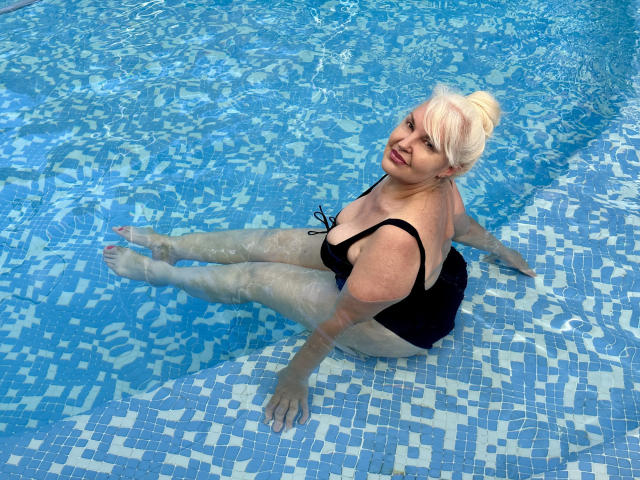 KamaliaSim with pale skin and blonde hair, wearing a black one-piece, sitting in a blue-tiled pool, legs out, smiling, water showing on her skin. Boobs visible.