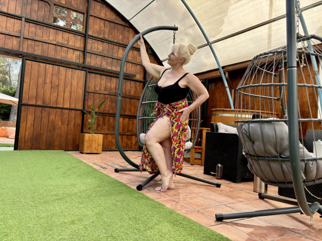 KamaliaSim with blonde hair, black top, and colorful floral skirt, standing barefoot on tile, holding hanging chair, showing her big ass and thick thighs, in a wooden outdoor area.