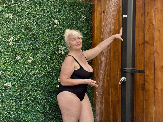 KamaliaSim stands in a shower, black one-piece, water pouring, right arm raised, blonde hair, medium tits, curvy, smiling, wood panel wall, green textured wall, water stream visible.
