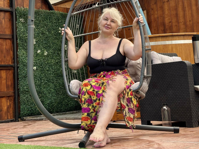 KamaliaSim with big tits, blonde hair, black bra, colorful skirt, sitting in a hanging chair, holding ropes, barefoot, wooden wall in background, smiling, relaxed.
