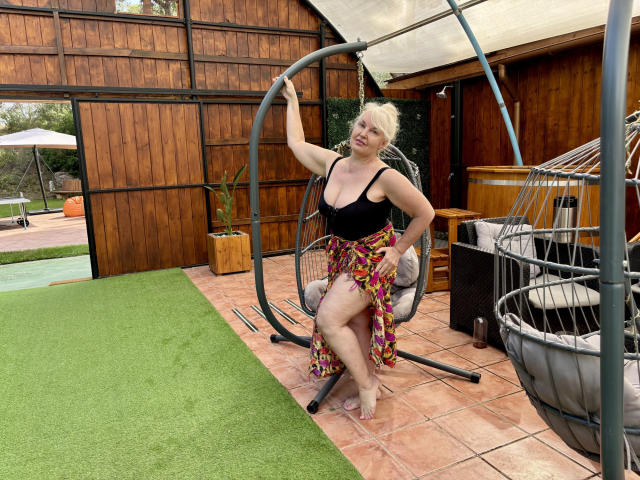 KamaliaSim with blonde hair, black top, and colorful skirt, sitting in a hanging chair, showing her tits and ass, hand on chair, on a tiled patio, green grass.