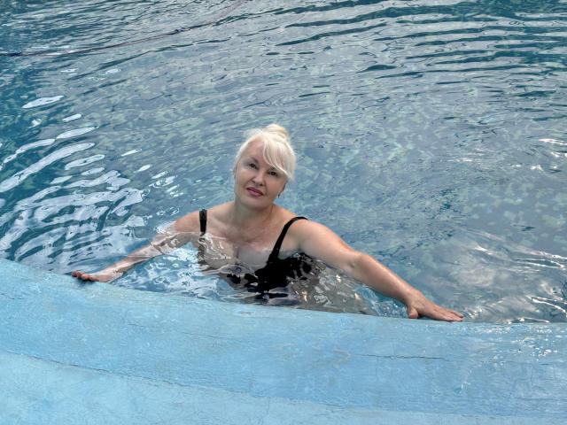 KamaliaSim with pale blonde hair, black one-piece swimsuit, and small tits, floating in a blue-tiled pool, arms resting on the edge, water rippling around her.