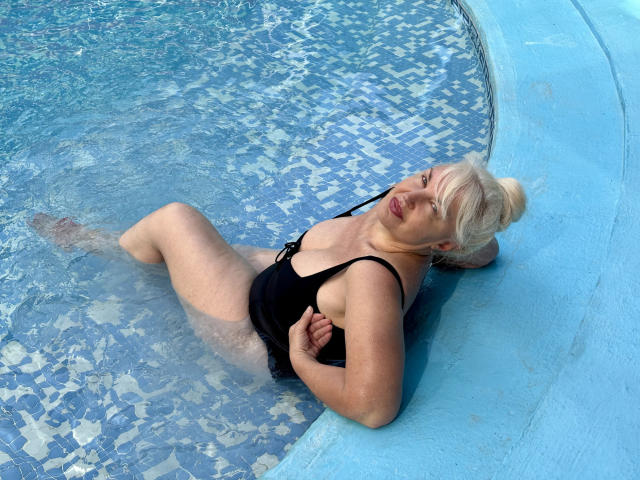 KamaliaSim with blonde hair, black one-piece swimsuit, medium tits, lying in blue-tiled pool, water up to her ass, arms behind head, eyes closed, relaxed.