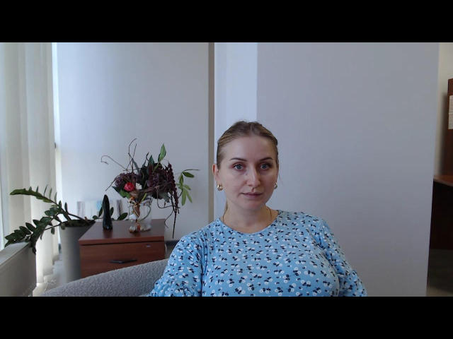Deriina with blonde hair in a blue patterned hospital gown, sitting, looking at camera, small tits, fair skin, wooden dresser with plants, white walls, window with curtains.