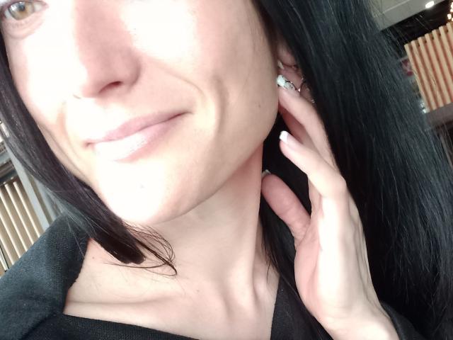 BellaJulls with black hair, pale skin, subtle smile, and light pink lips, touching her ear with a hand, wearing a black top, in a bright room.
