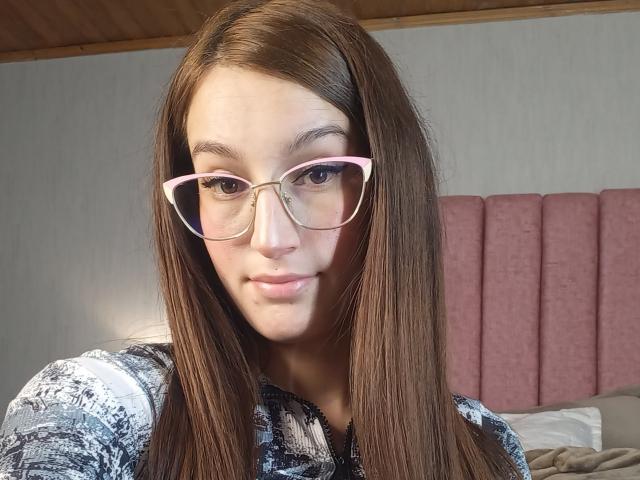 RitaJonness with long brown hair, pink-rimmed glasses, and a subtle smile, wearing a grey and black patterned top, showing her cute face and small, pink lips.
