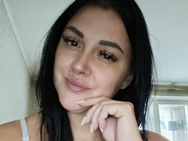 XCrazyBabeX has dark hair, thick eyebrows, long eyelashes, glossy pink lips, and a cute smile. She's resting her chin on her hand with white nails.