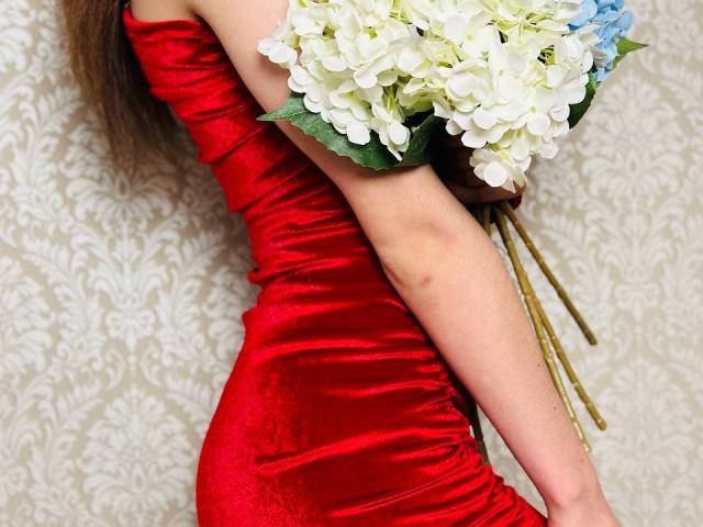 Harisma, vestita con un elegante abito rosso di velluto, tiene un bouquet di ortensie bianche e blu, con un braccio e alcune foglie verdi visibili, e i suoi capelli castani che le scendono lungo la schiena..