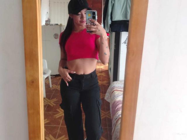 SashaHans indossa una canotta rossa che mette in mostra i suoi addominali, pantaloni neri e un berretto nero, sta scattando un selfie allo specchio, tiene in mano un telefono, ha dei tatuaggi sul braccio, si trova in una camera da letto..