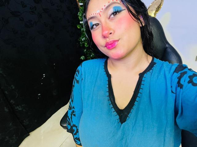 KalpanaAisha has pink pouty lips, blue eye makeup, and a red dot pattern on her forehead. She's wearing a blue top with black trim, looking at the camera.
