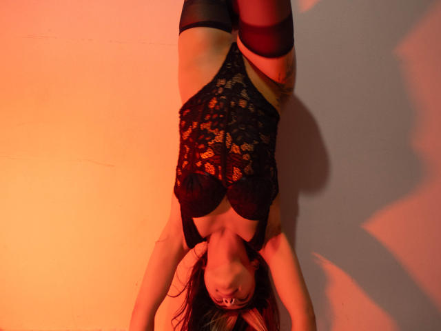 MandyCuteNSexy is upside down, wearing a black lace bodysuit, red stockings, and has large tits. She's laying on a flat surface with orange lighting.