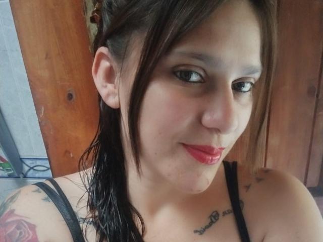 MaiteRM has a side-swept wet brown ponytail, red lipstick, black tank top, tattoo on her right shoulder, and a visible tattoo under her left collarbone.