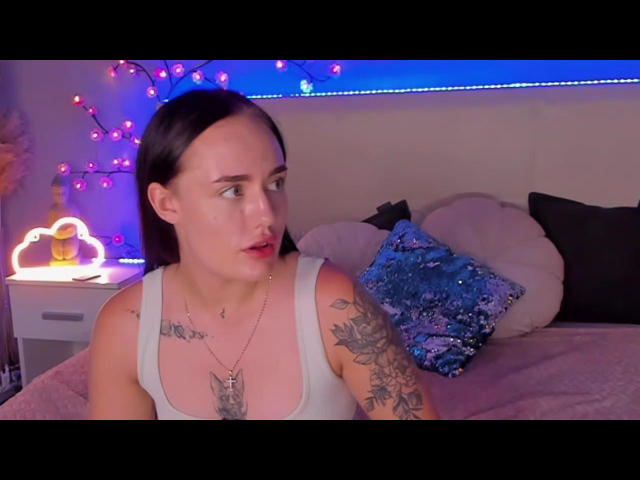 SaraFlame met donker haar, een wit tanktop, tatoeages op de rechterarm en borst, die naar de zijkant kijkt, rode lippenstift, een blauw decoratief kussen op een roze bed, paarse lichtjes..