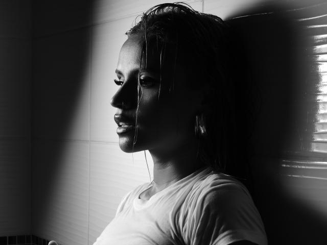 Black-and-white photo of MartinaNovac with wet hair, white tee, standing in shadow, light highlighting her profile, showing her small tits and serious expression.