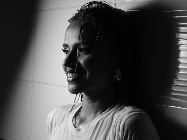Black and white photo of MartinaNovac with wet, dark hair, smiling, wearing a white shirt, standing against a wall with light and shadow on her right side.
