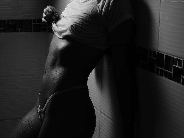 MartinaNovac lifting a shirt, revealing a wet, toned, muscular torso and wearing a thin, white thong, with dark tiles in the background. Her skin is glistening.