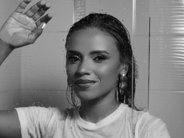 Black-and-white photo of MartinaNovac with wet, slicked-back hair, wearing a white shirt, standing in a shower with water droplets on the glass. She's smiling and waving.