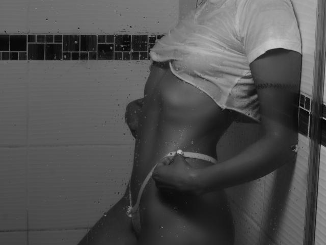 Black-and-white photo of MartinaNovac with wet white shirt, exposing small tits and slim, toned body, pulling down thong, showing her bare ass and a tattoo on her arm.
