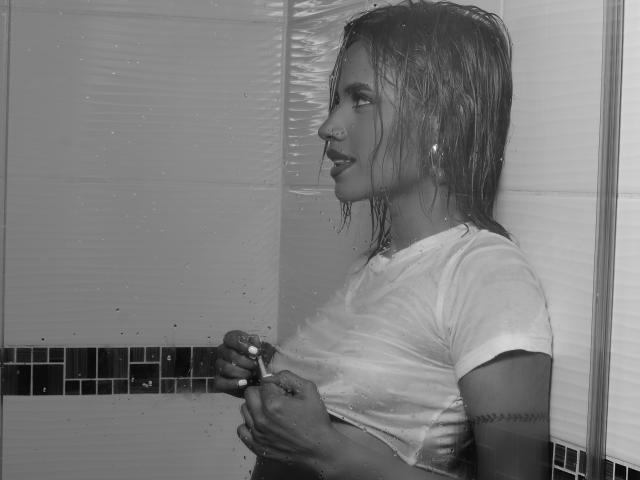 Black-and-white photo of MartinaNovac with wet, shoulder-length hair, wearing a soaked white t-shirt, showing small tits, holding shirt, tattoo on left arm, in a shower.