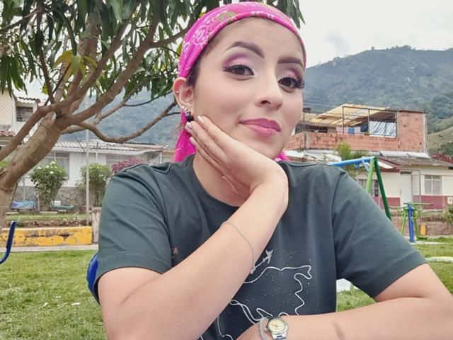 LizBonnet with pink headscarf, dark green tee, pink lipstick, and hand on cheek, smiling, showing off her titty ring, in front of a tree, wearing a watch.