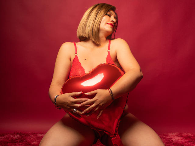 JolieDuvalle met kort blond haar, rode kant lingerie, die een glanzend rood hart vasthoudt, zittend met gespreide benen, rode achtergrond, lachend met gesloten ogen, en armbanden draagend..