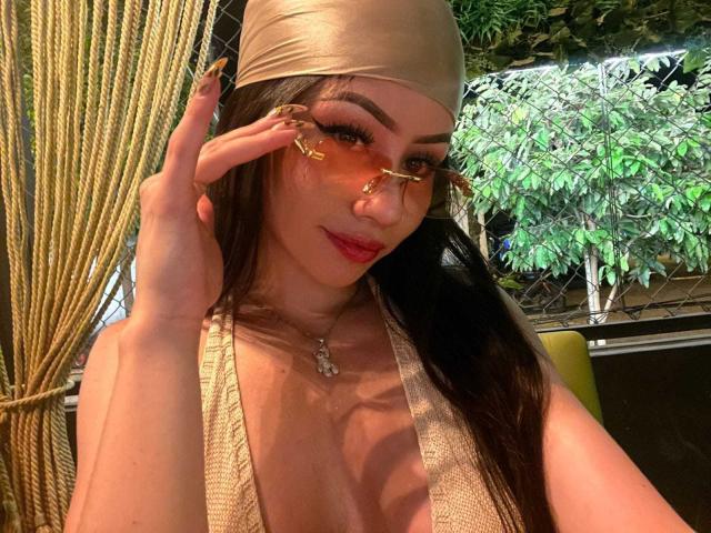 KattRouse with gold headwrap, orange-tinted glasses, and long nails, showing cleavage in a sheer, gold top, smiling with red lipstick, in a dimly-lit, green-leafed environment.